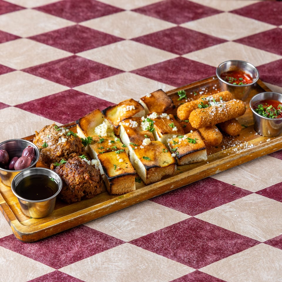 Wooden platter of grilled bread, croquettes, meatballs, olives, and dipping sauces on a red-and-white checkered tablecloth.
