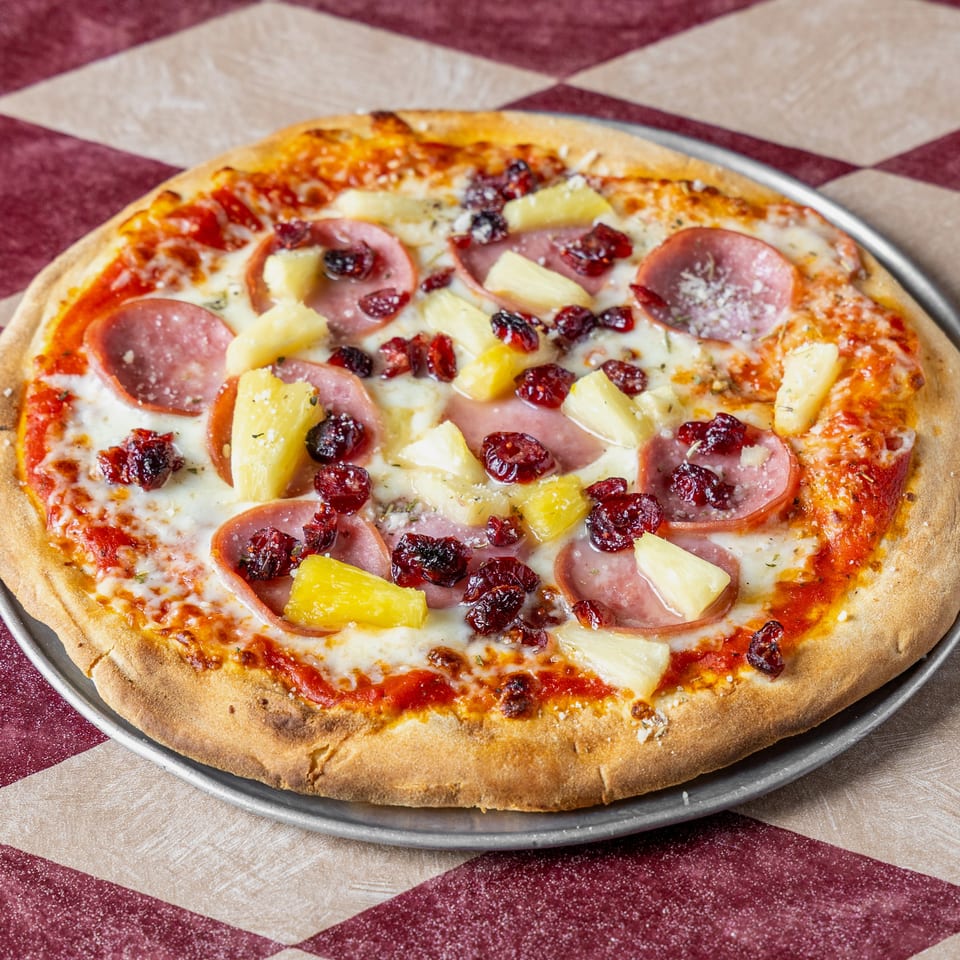Cheesy pizza topped with sliced sausage, pineapple chunks, and dried cranberries on a metal tray over a checkered tablecloth.