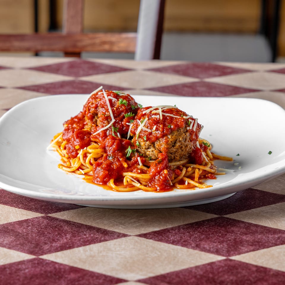 Spaghetti topped with tomato sauce, meatballs, shredded cheese, and parsley on a white plate atop a checkered tablecloth.