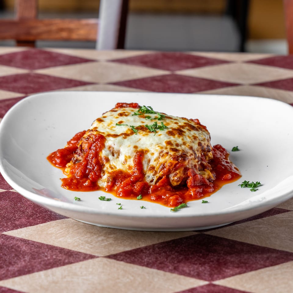 Melted cheese-covered entrée served with tomato sauce and parsley on a white plate atop a burgundy checkered tablecloth.